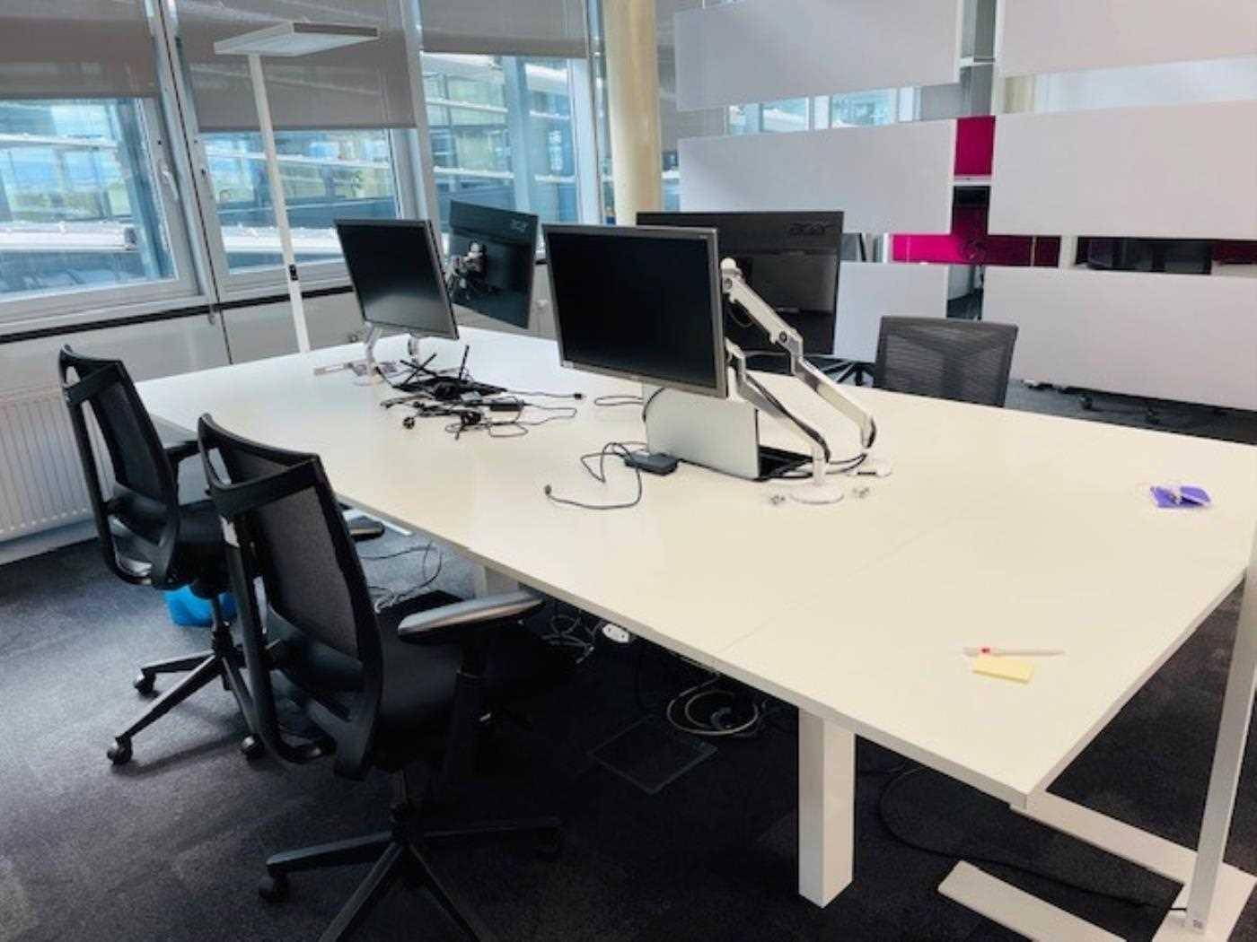 Moderner Bürotisch mit mehreren Monitoren und ergonomischen Stühlen, Blick auf das Gebäudeinnere.
