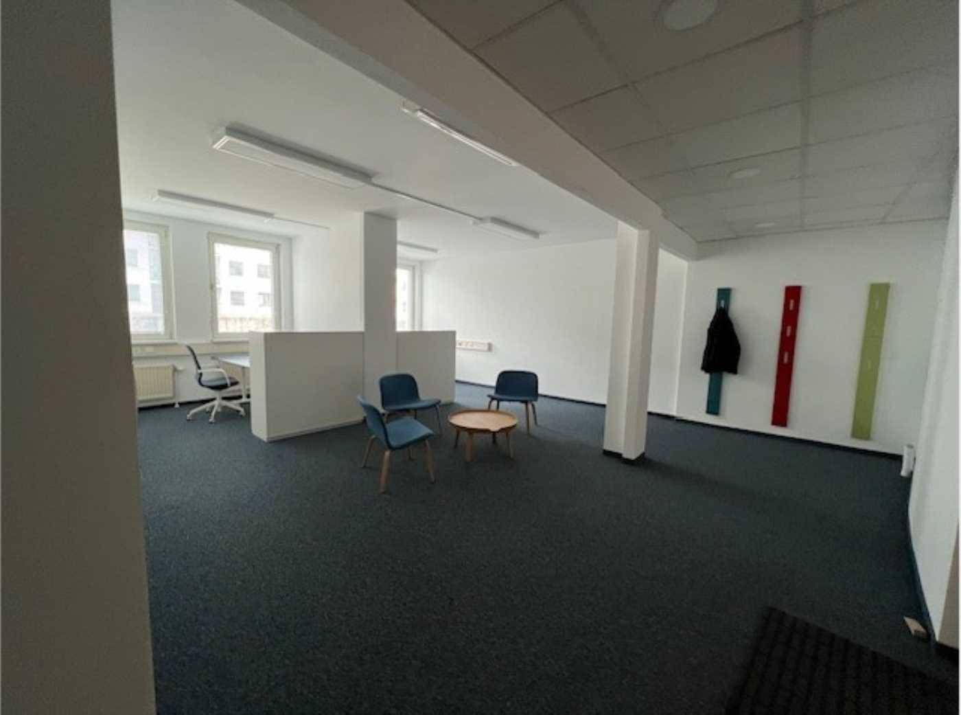 Heller Büroraum mit Teppichboden, weißen Wänden und einer kleinen Sitzecke.