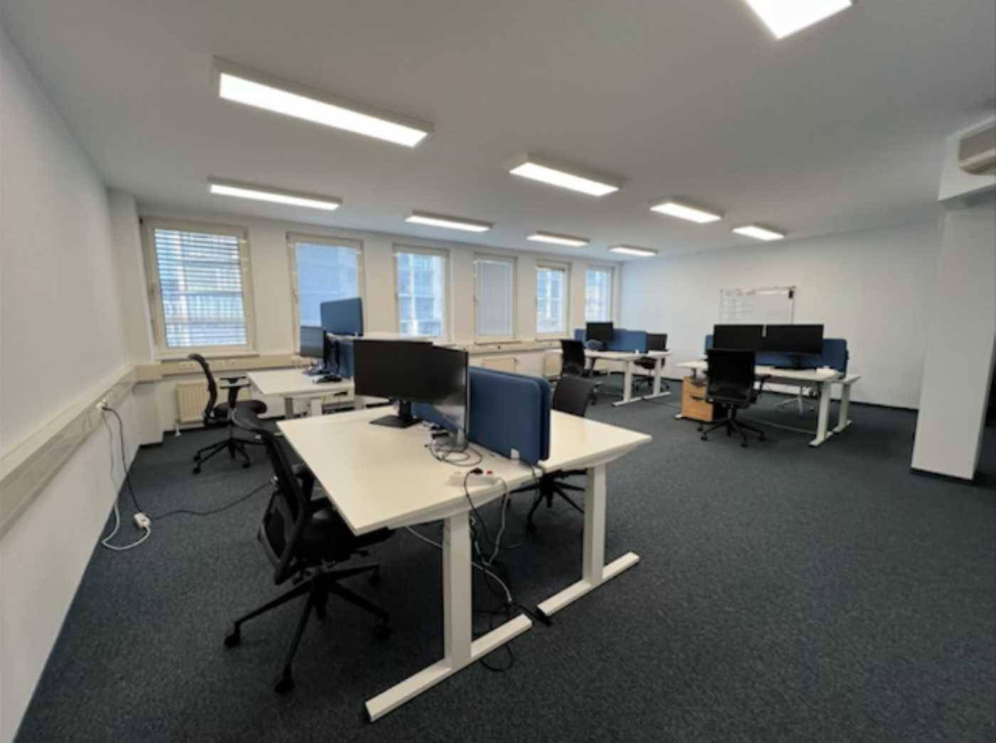 Großraumbüro mit mehreren Arbeitsplätzen, Computern und Fenstern mit Jalousien.