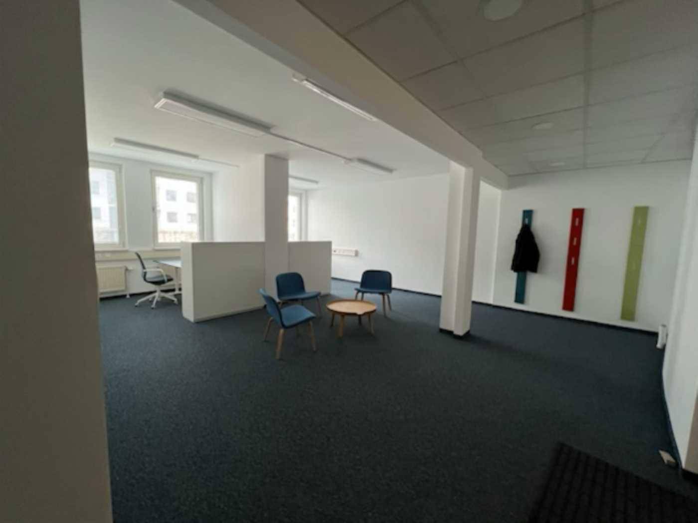 Heller Büroraum mit Teppichboden, weißen Wänden und einer kleinen Sitzecke.