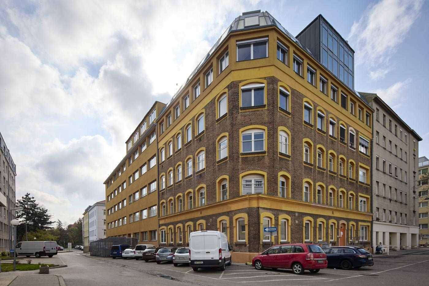 Großes Eckgebäude mit gelb-brauner Ziegelfassade und vielen Fenstern, umgeben von parkenden Autos.
