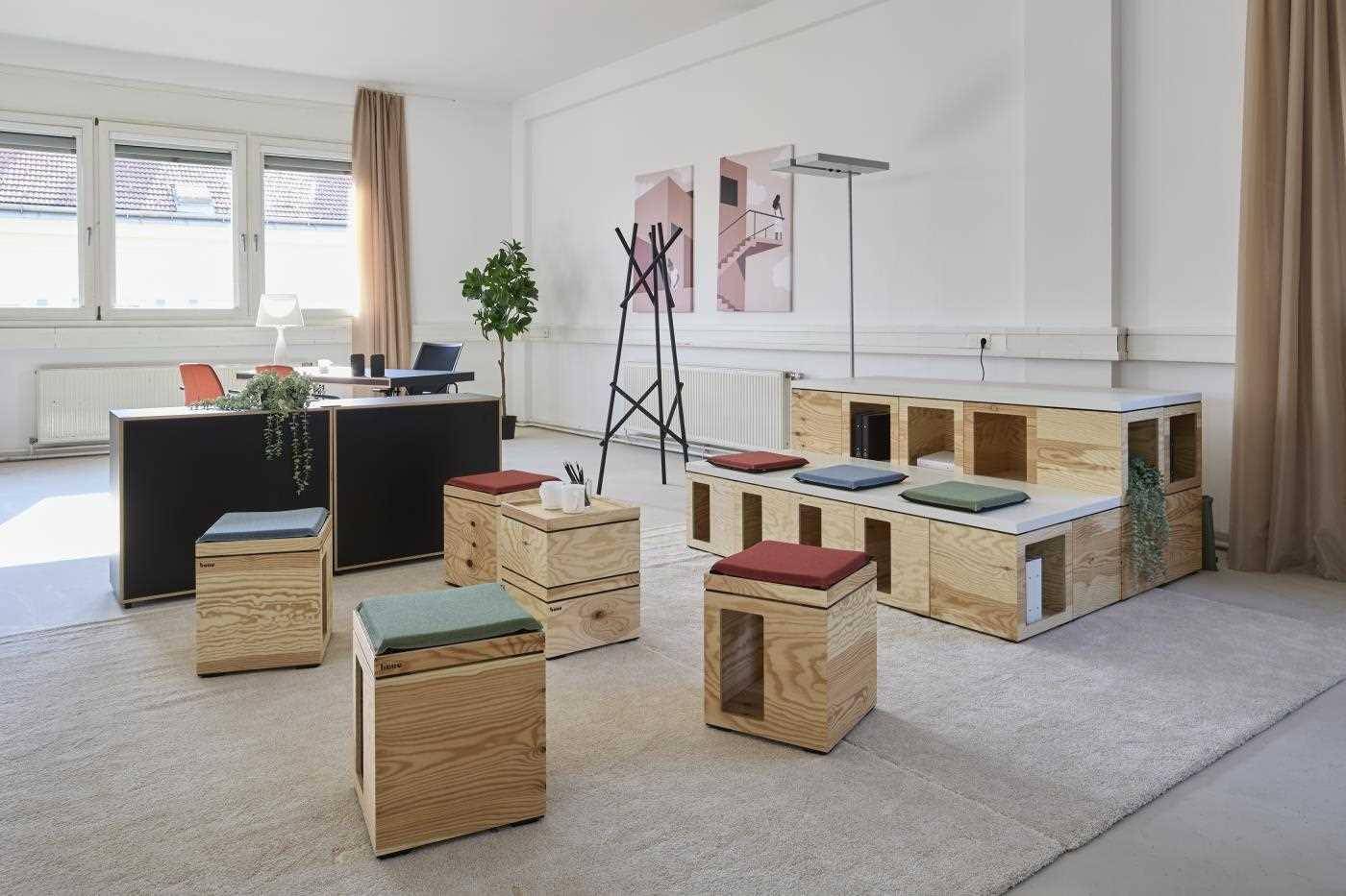 Heller, offener Büroraum mit großen Fenstern, modularen Holzmöbeln und einem Schreibtisch im Hintergrund.