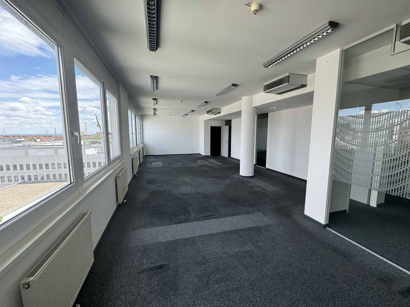 Langer Büroraum mit durchgehender Fensterfront, Teppichboden und Säulen, bietet einen weiten Stadtblick.