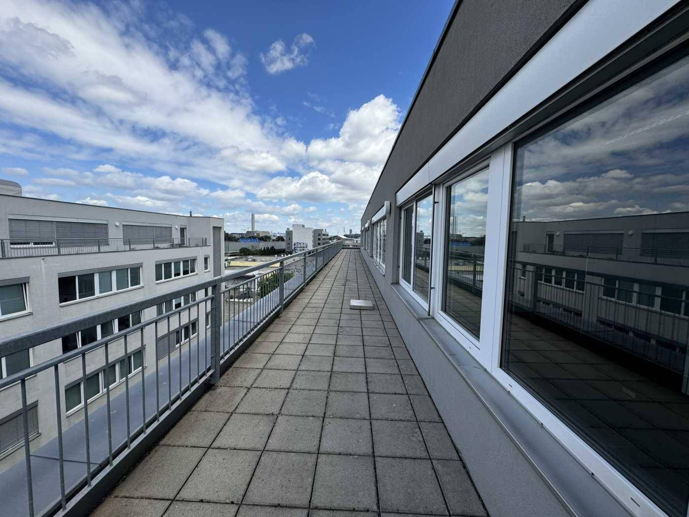 Lange Außenterrasse mit Fliesenboden und Metallgeländer, bietet einen weiten Blick über die umliegenden Gebäude.