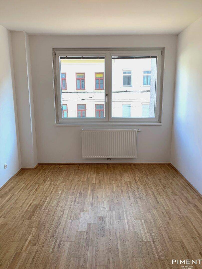 Leerer Raum mit Holzboden und einem großen Fenster mit Blick auf gegenüberliegende Gebäude.