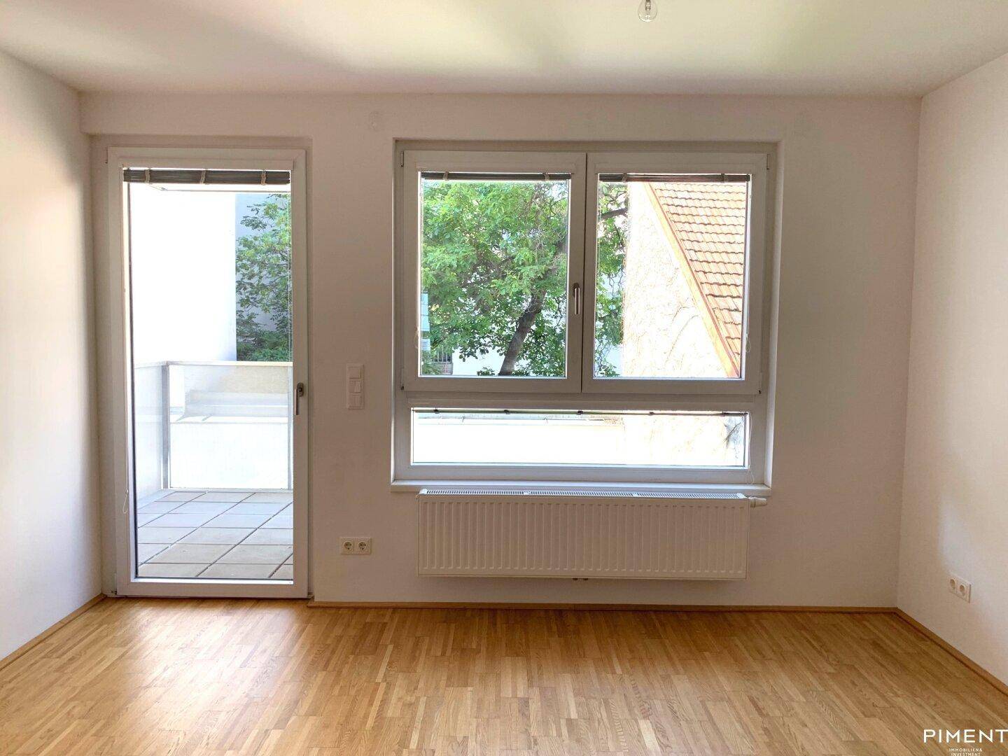 Heller Raum mit Holzboden, großem Fenster und Zugang zu einem Balkon mit grüner Aussicht.