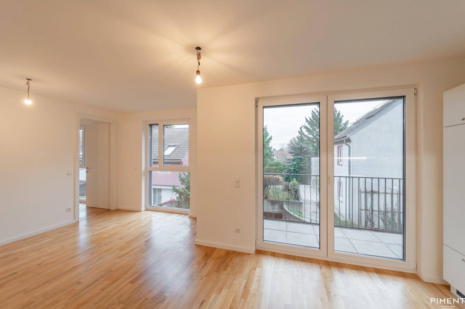 Geräumiger Wohnbereich mit Holzboden, großen Fenstern und Zugang zu einem Balkon.