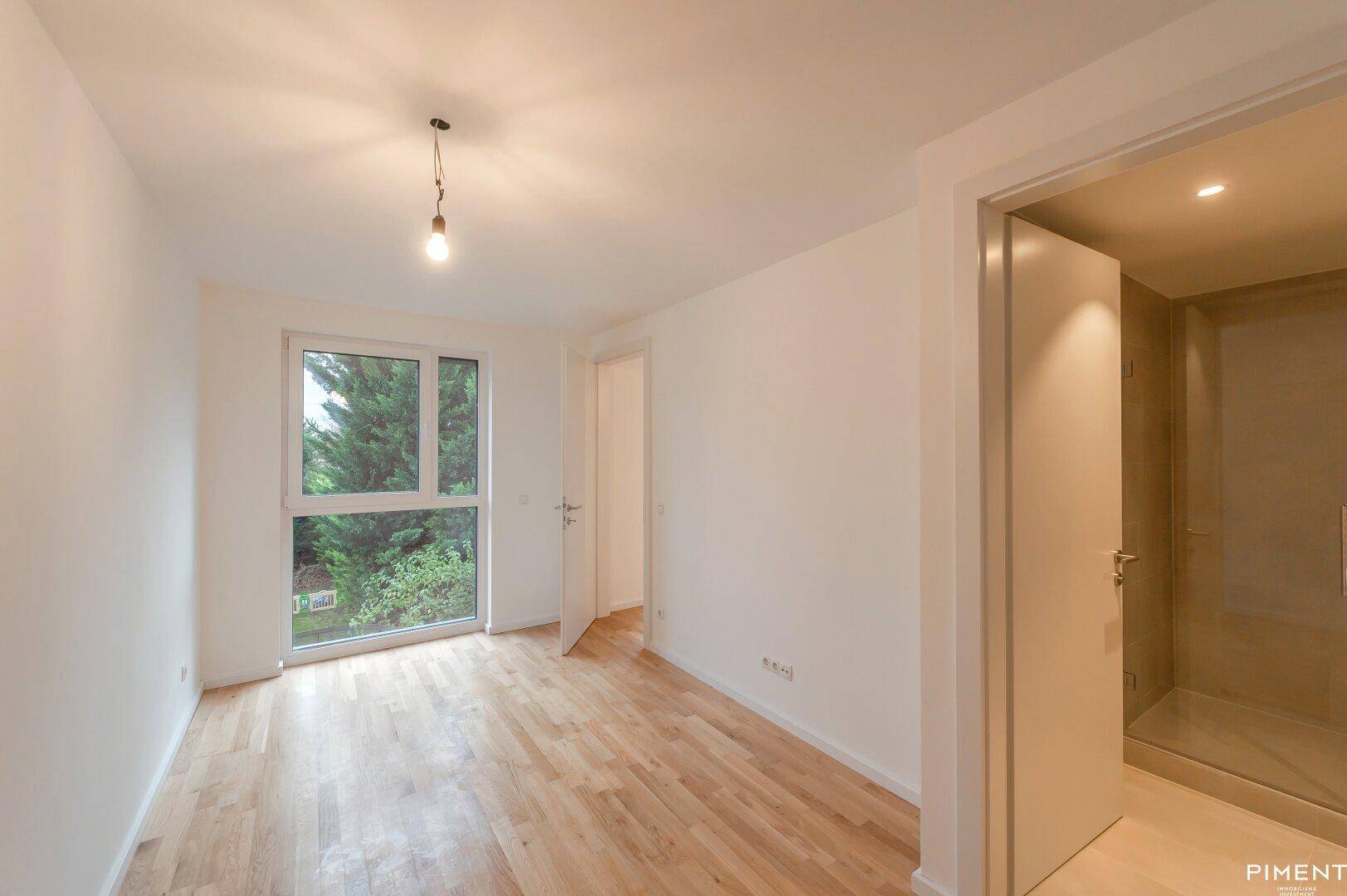 Helles Schlafzimmer mit Parkettboden und großem Fenster, angrenzend an ein Badezimmer.