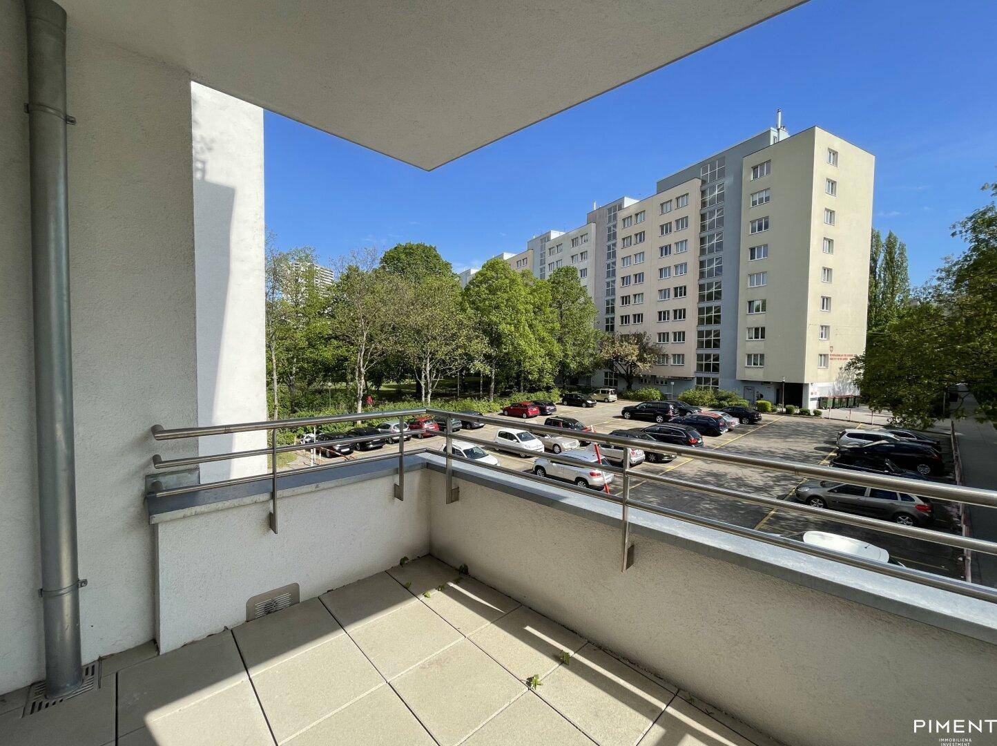 Sonniger Balkon mit Fliesenboden und weitem Blick auf die Wohnanlage und Bäume.