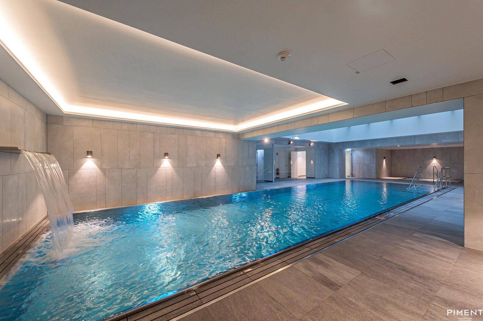 Modernes Hallenbad mit eleganten Fliesen, integriertem Wasserfall und stimmungsvoller Beleuchtung.
