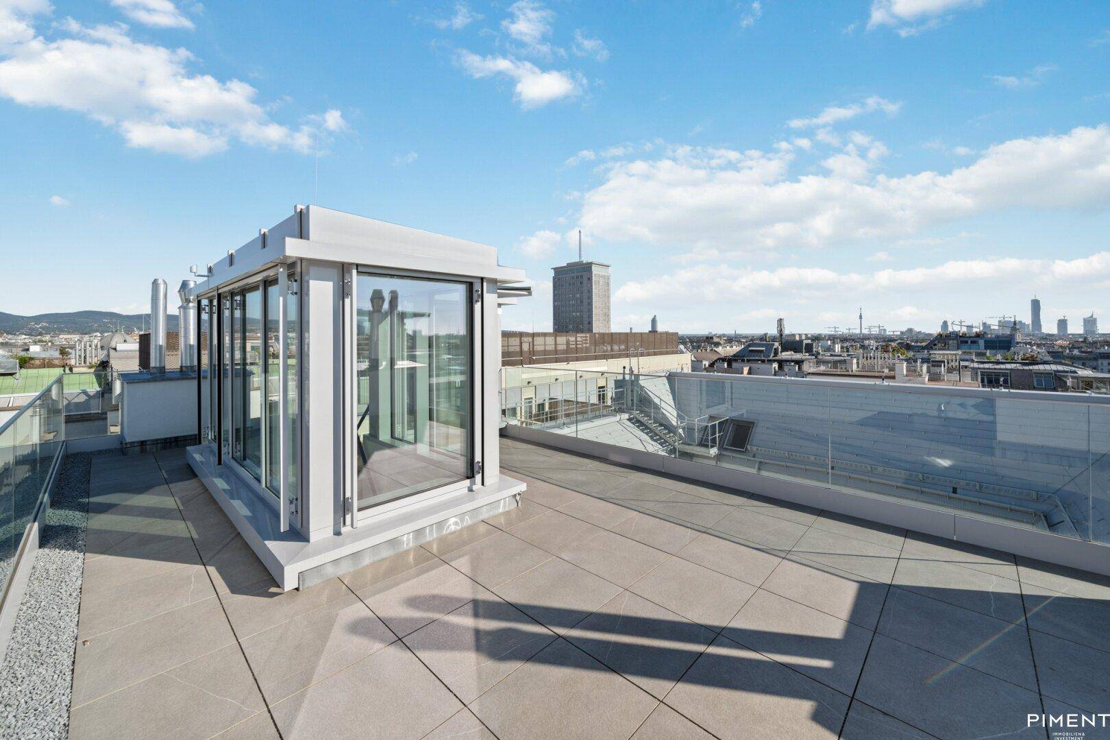 Geräumige Dachterrasse mit moderner Glasstruktur und weitem Stadtblick unter blauem Himmel.