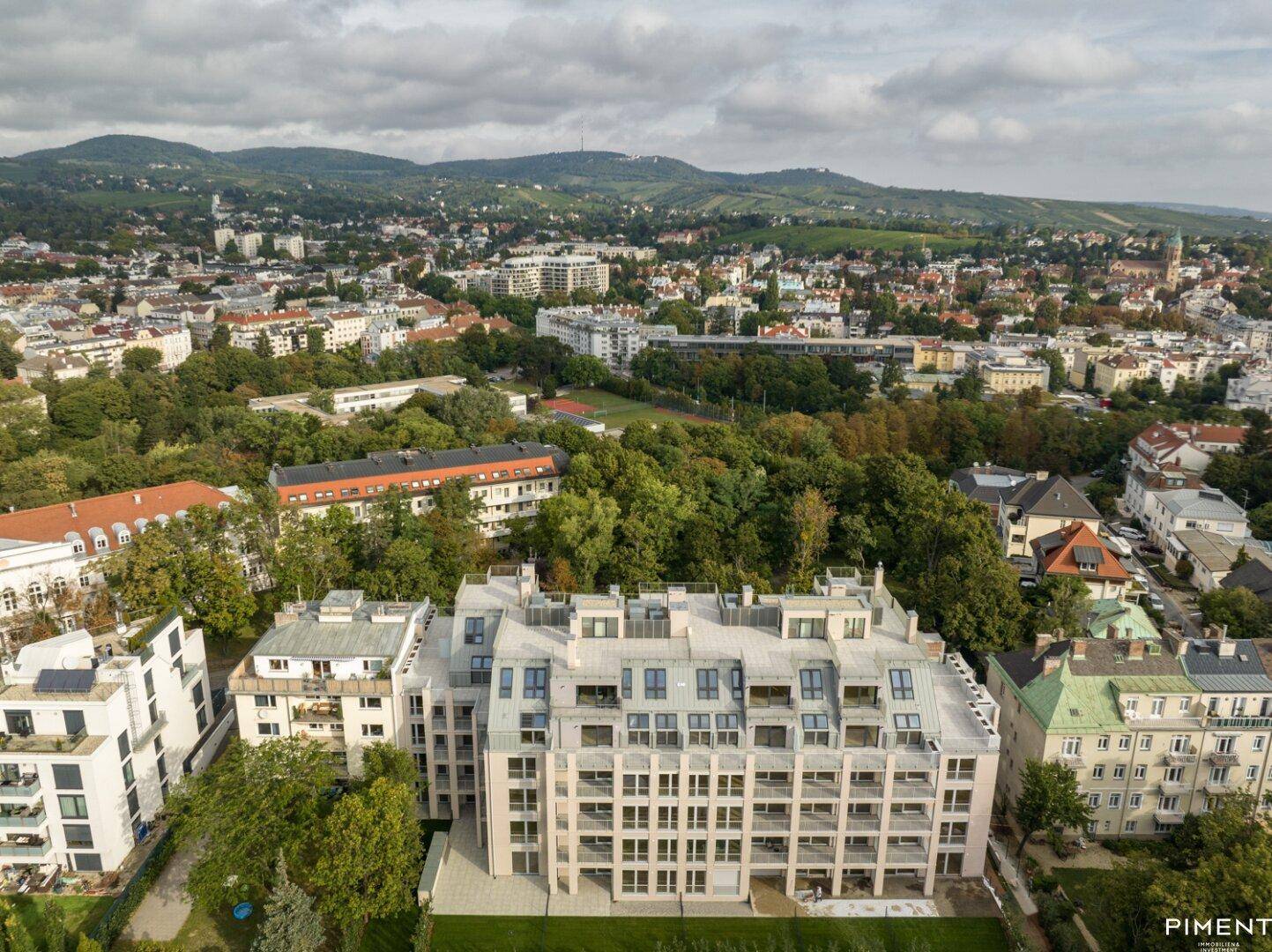 Luftaufnahme eines modernen Wohnkomplexes, umgeben von viel Grün und Hügeln mit Weinbergen.