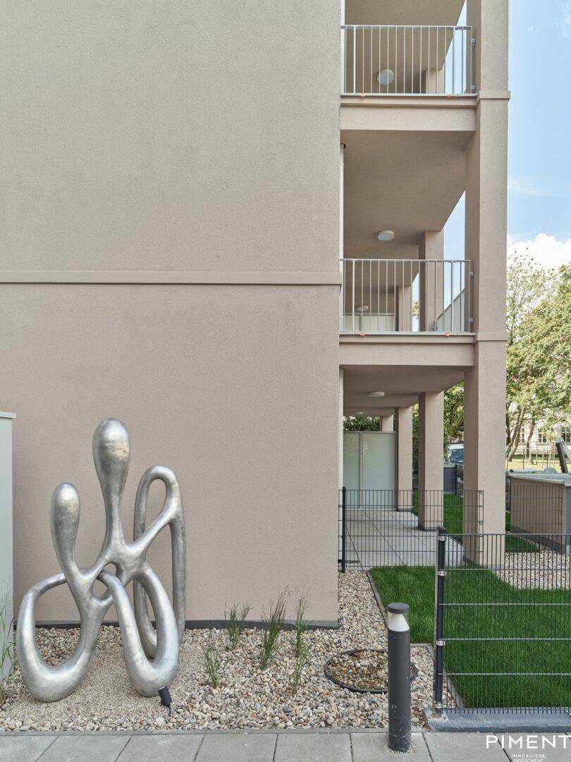 Moderne Gebäudefassade mit Balkonen und dekorativer Skulptur im Vorgartenbereich.