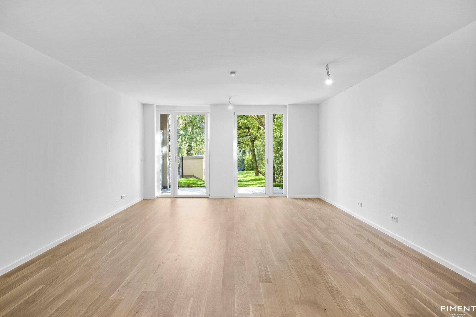 Heller Wohnbereich mit Holzboden und zwei großen Fenstertüren, die ins Grüne führen.