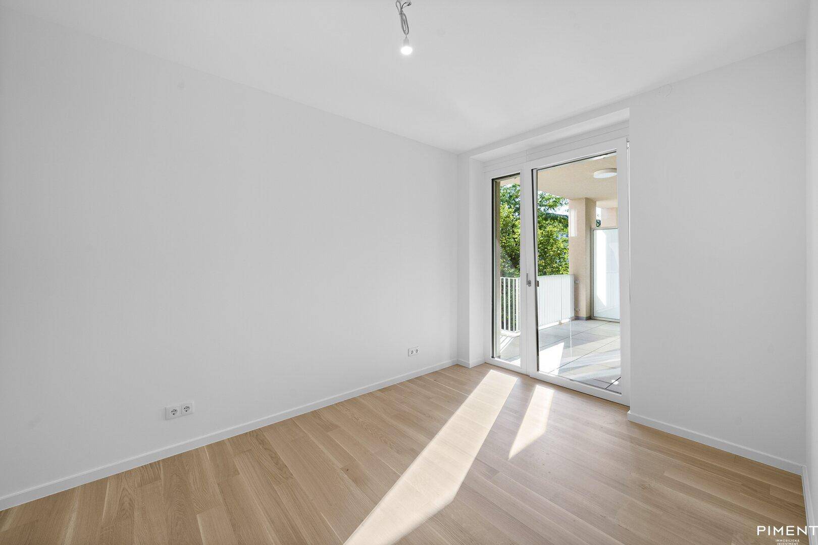 Leeres Schlafzimmer mit hellem Parkettboden und direktem Zugang zu einem sonnigen Außenbereich.