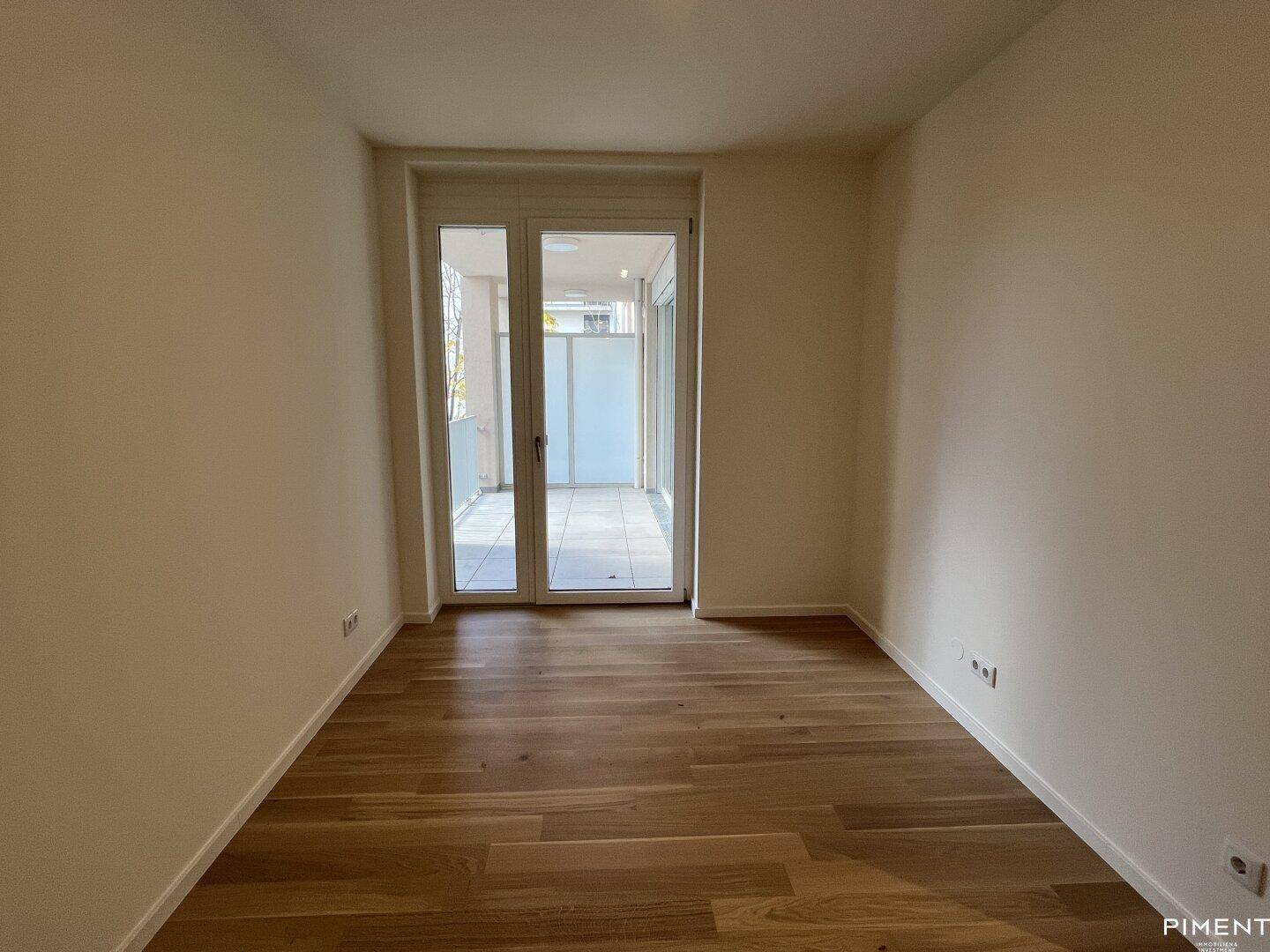 Heller Raum mit Parkettboden und großem Fenster, das Zugang zu einem Balkon bietet.