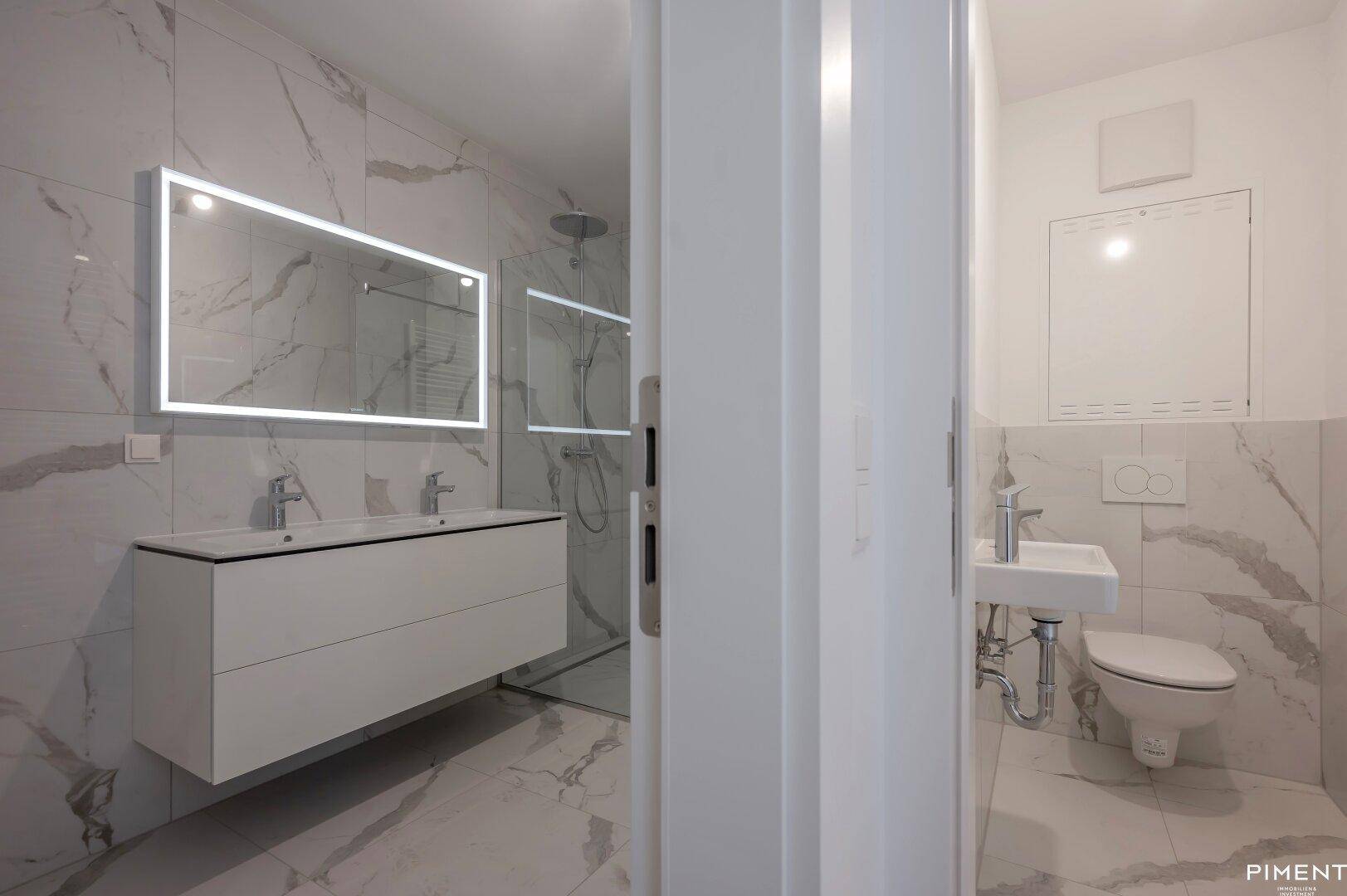 Modernes Badezimmer mit Marmoroptik-Fliesen, Doppelwaschbecken und beleuchtetem Spiegel, angrenzend an ein separates WC.