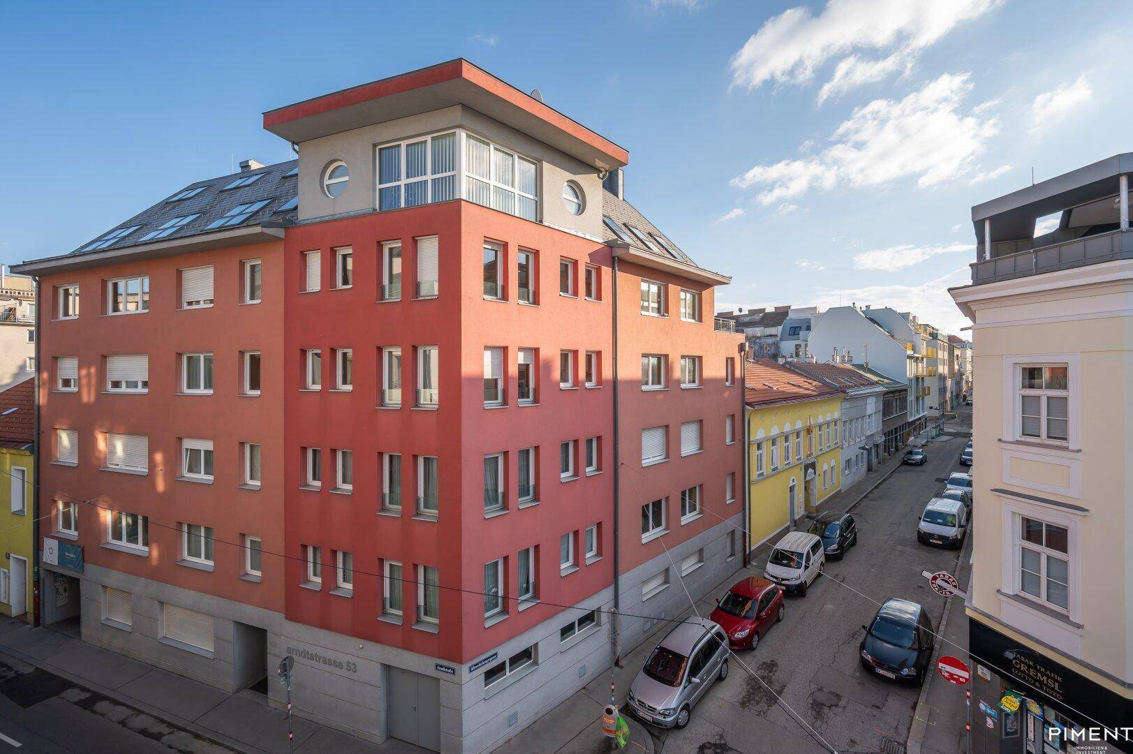 Rotes Mehrfamilienhaus mit zahlreichen Fenstern an einer städtischen Straße unter blauem Himmel.