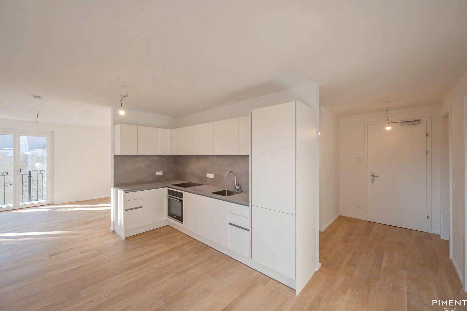 Moderne Einbauküche mit weißen Schränken, grauer Arbeitsplatte und integriertem Backofen in einem offenen Wohnbereich.