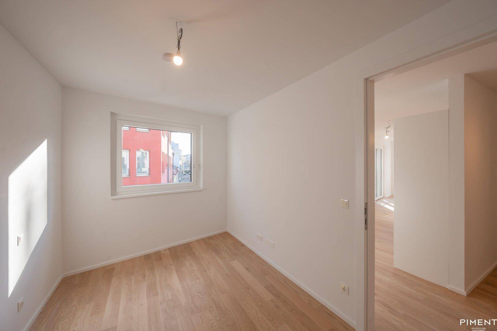 Helles Zimmer mit Parkettboden und weißen Wänden, bietet einen Blick auf die gegenüberliegende rote Fassade.