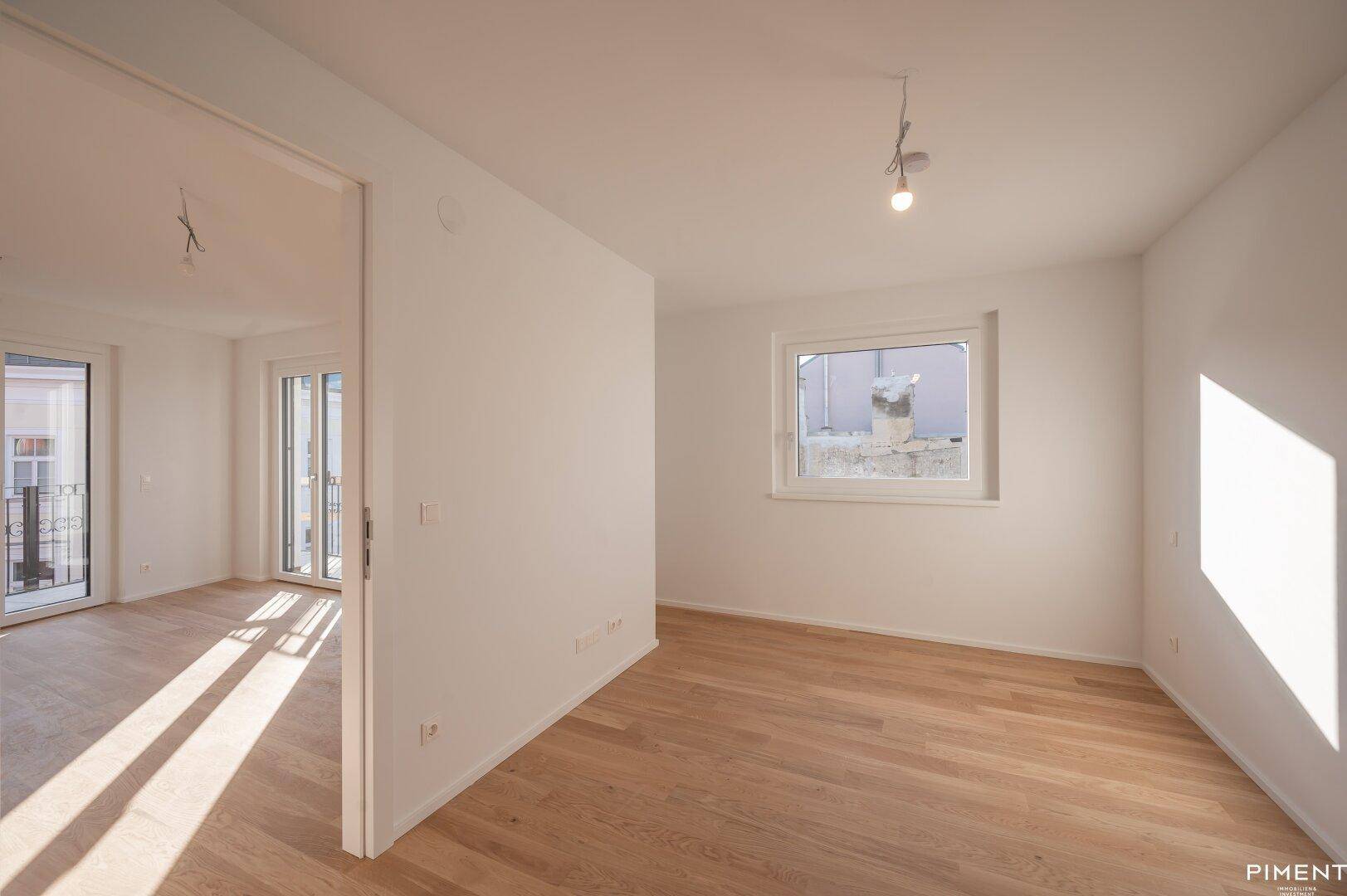 Leeres Zimmer mit hellem Parkettboden, weißen Wänden und einem Fenster, das viel Tageslicht hereinlässt.