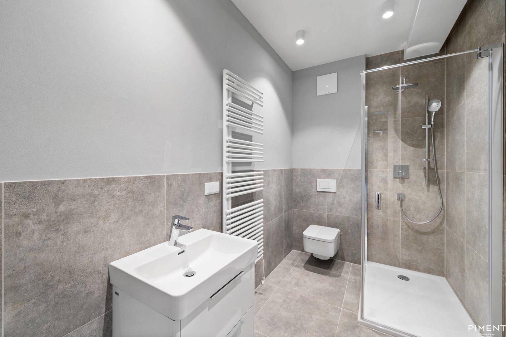 Modernes Badezimmer mit grauen Fliesen, Duschkabine, wandhängendem WC und Handtuchheizkörper.