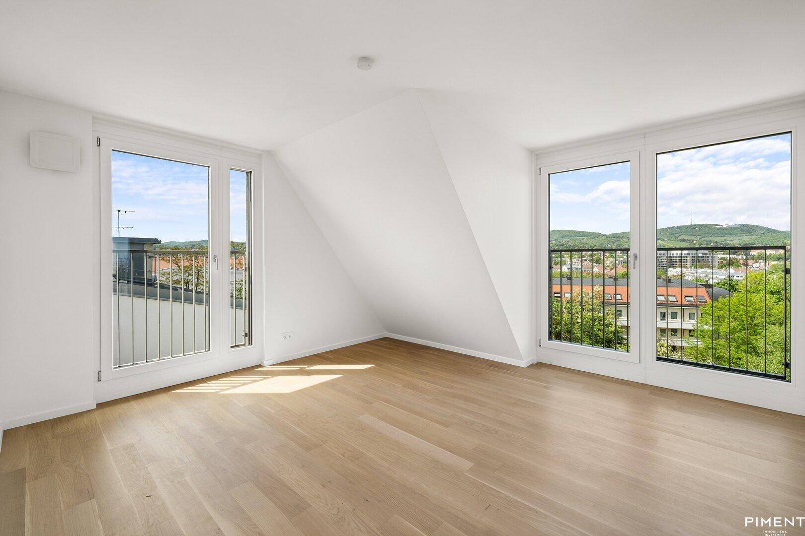 Lichtdurchflutetes Dachzimmer mit Holzboden und zwei großen Fenstern mit Stadtblick.