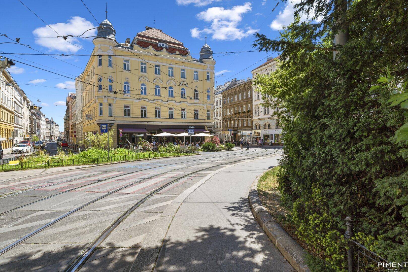 Belebte Straßenszene mit einem markanten gelben Gebäude, Straßenbahnschienen und Außengastronomie.