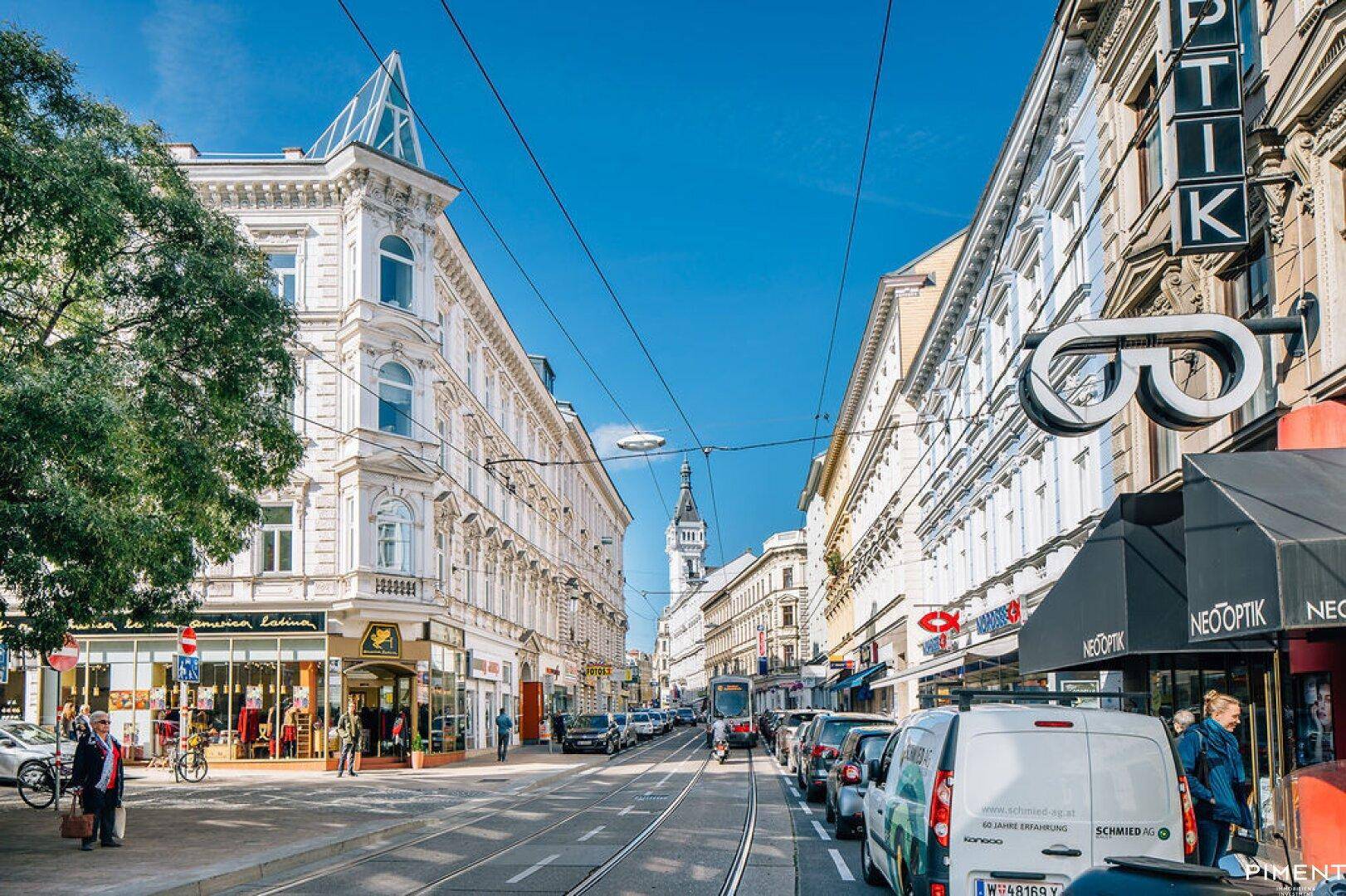 Belebte Stadtstraße mit historischen Gebäuden, Geschäften und regem Verkehr unter blauem Himmel.