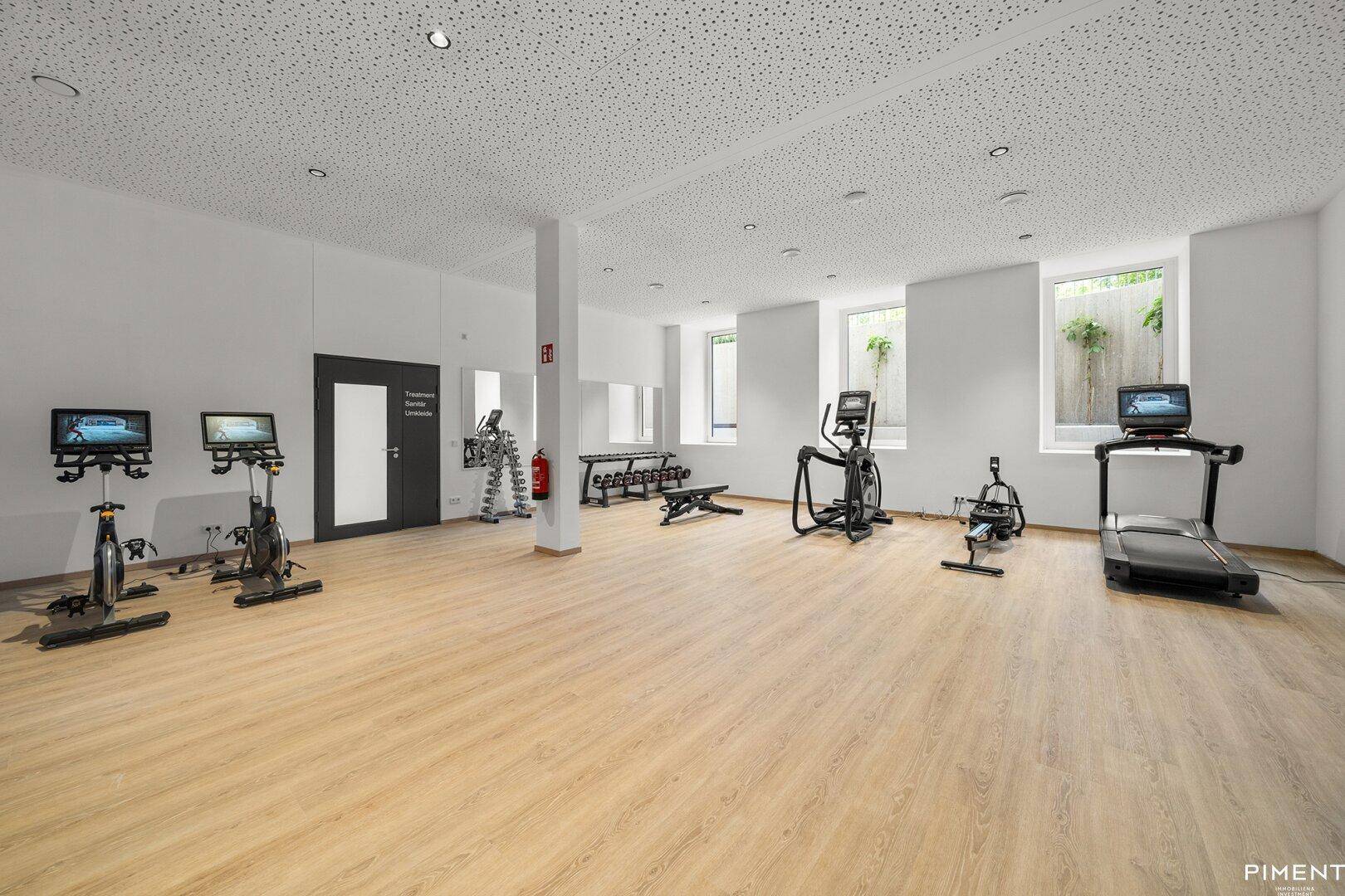 Geräumiger Fitnessraum mit verschiedenen Trainingsgeräten, großen Fenstern und hellem Holzboden.