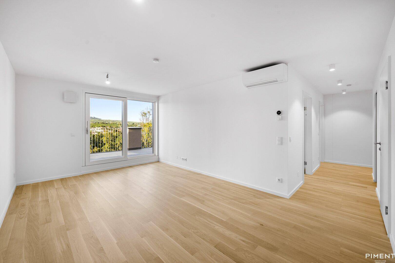 Helles, leeres Wohnzimmer mit Holzboden, großen Fenstern und Zugang zum Balkon.