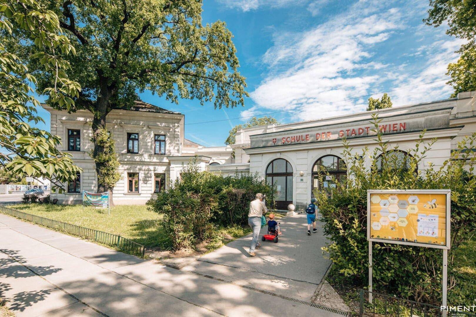 Historisches Schulgebäude der Stadt Wien mit gepflegten Grünflächen und einem sonnigen Eingangsbereich.