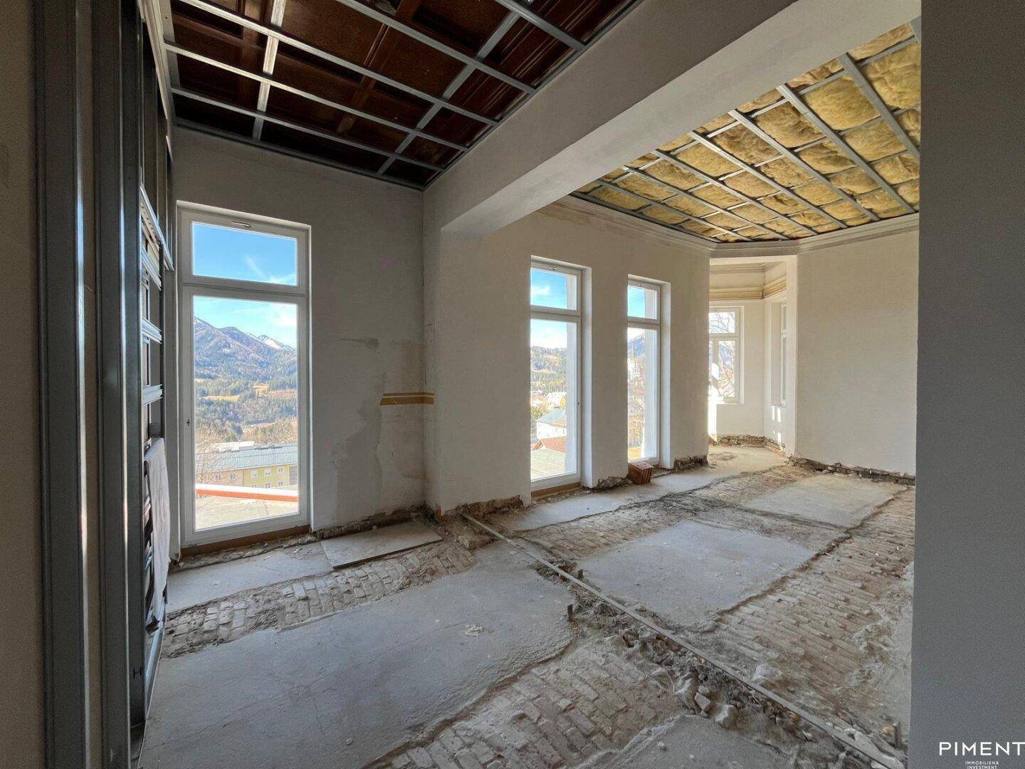 Innenraum im Rohbau mit hohen Fenstern und Blick auf die umliegende Landschaft.