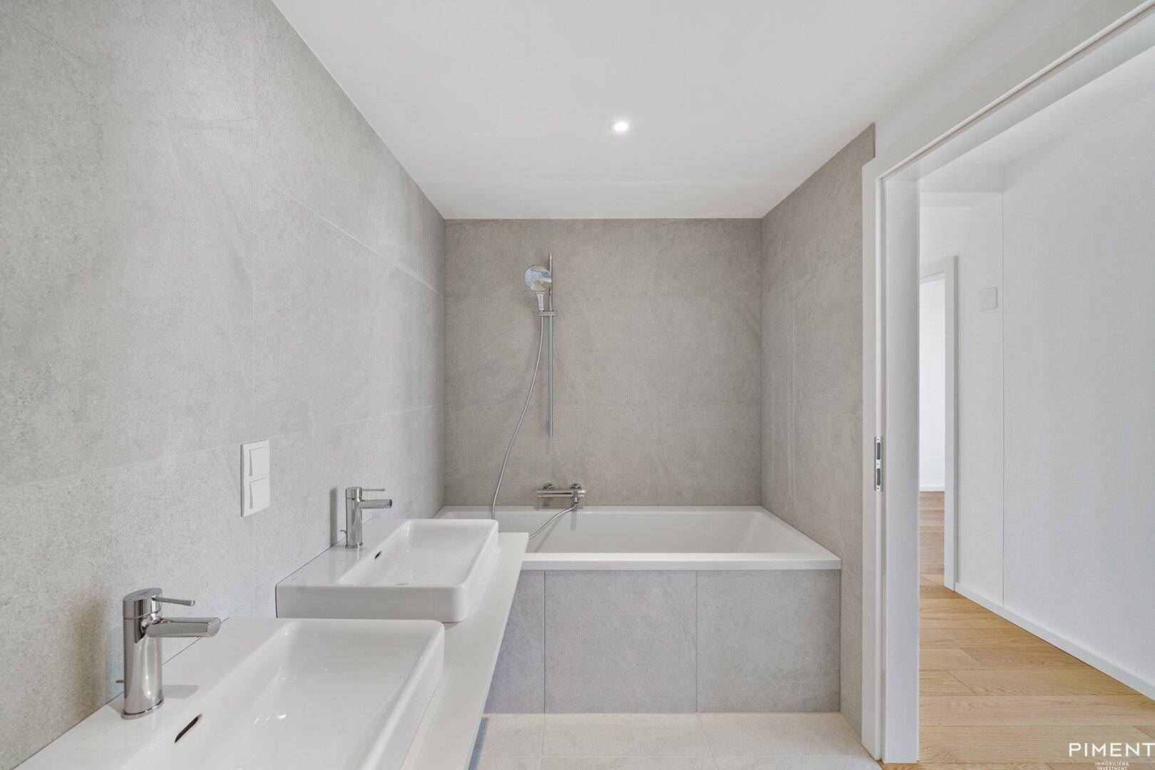 Elegantes Badezimmer mit Badewanne, Doppelwaschbecken und modernen grauen Fliesen an den Wänden.