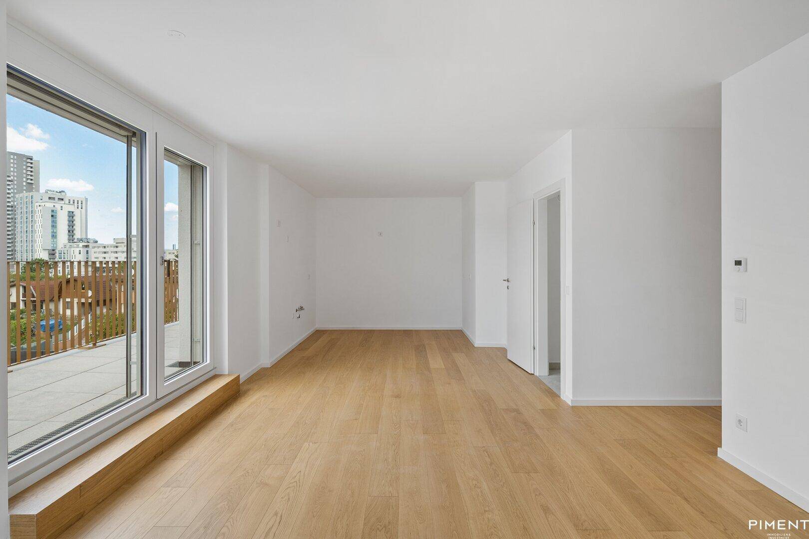 Helles, leeres Wohnzimmer mit großen Fenstern, Holzboden und Zugang zu einer Terrasse.