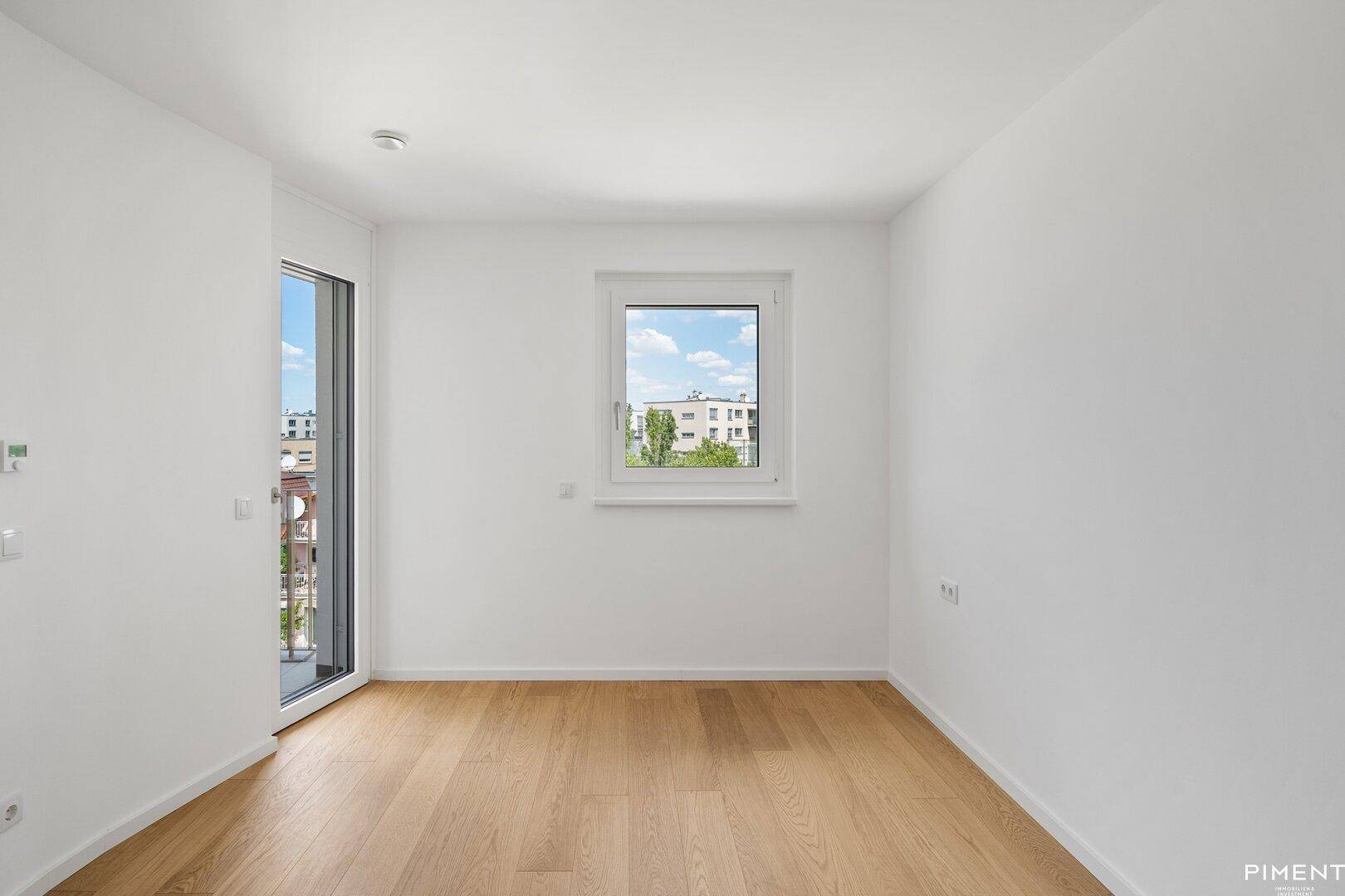 Helles Schlafzimmer mit Parkettboden und Fenster sowie Zugang zu einem kleinen Balkon.