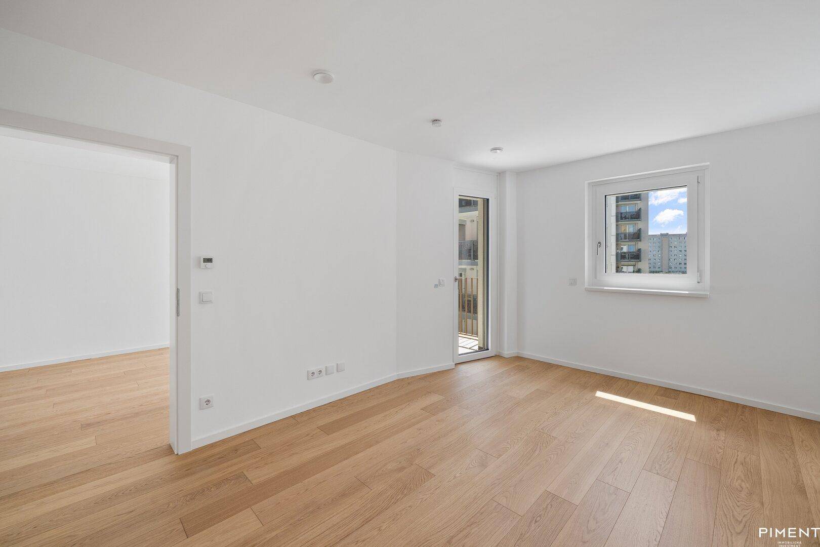 Helles Zimmer mit Eichenparkett, weißen Wänden und Zugang zu einem kleinen Balkon oder einer Terrasse.