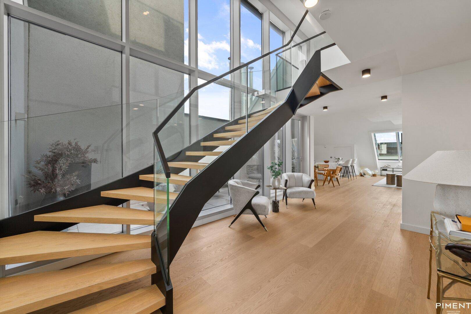 Elegante Holztreppe mit Glasgeländer und großen Fenstern, die viel Licht hereinlassen.