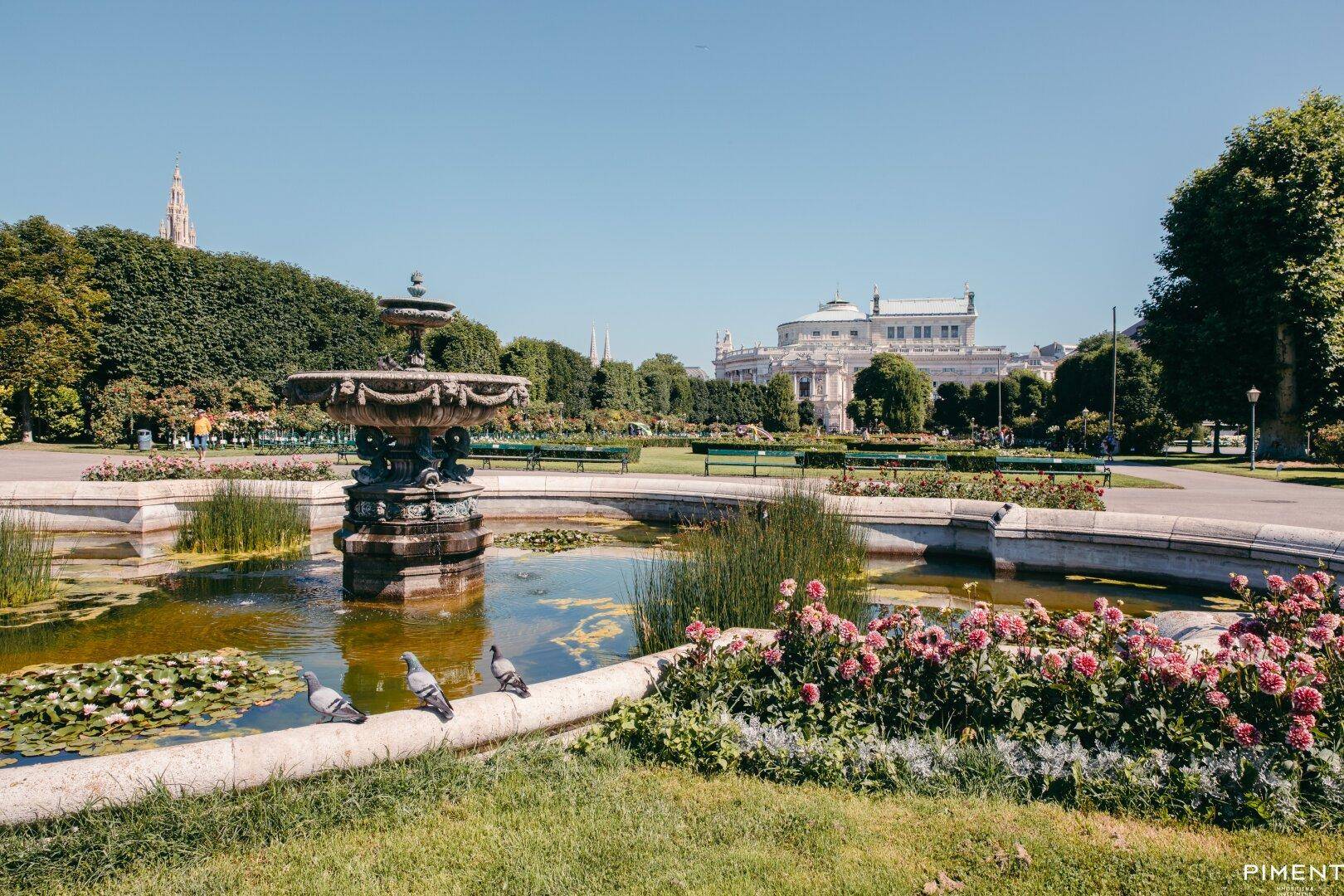 Malerischer Park mit einem großen Brunnen, blühenden Blumenbeeten und üppigem Grün.