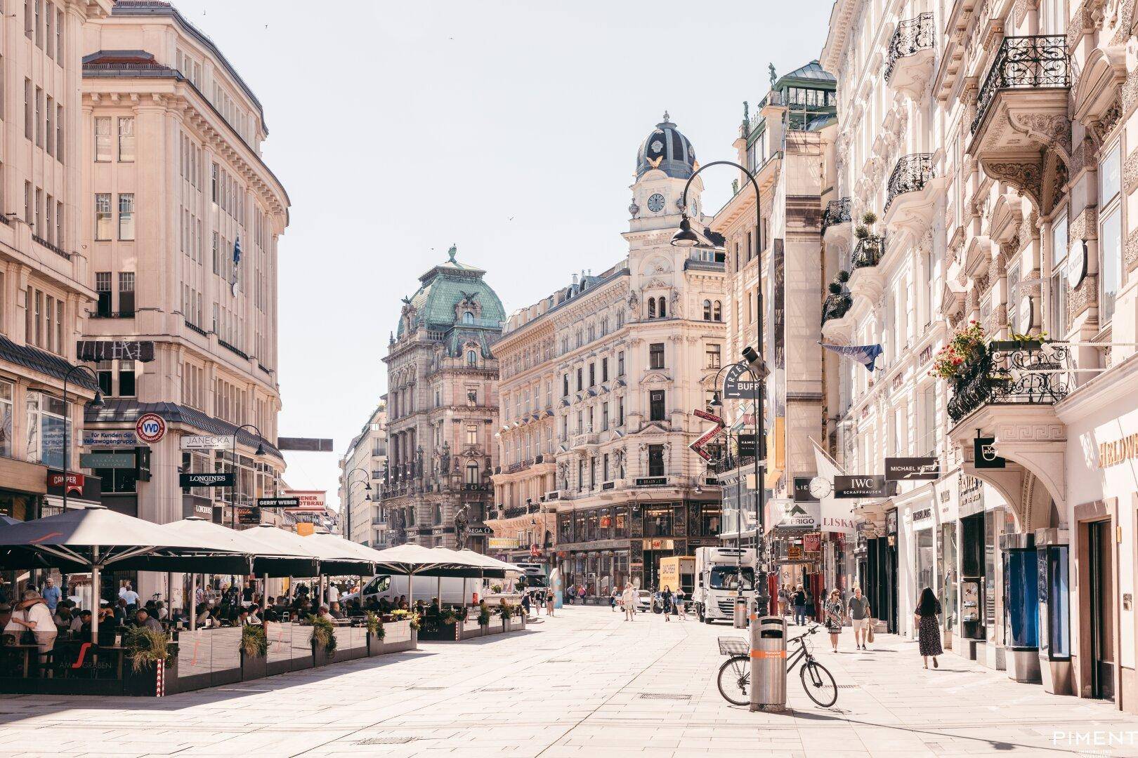 Belebte Einkaufsstraße mit historischen Gebäuden, Geschäften und Außengastronomie unter blauem Himmel.