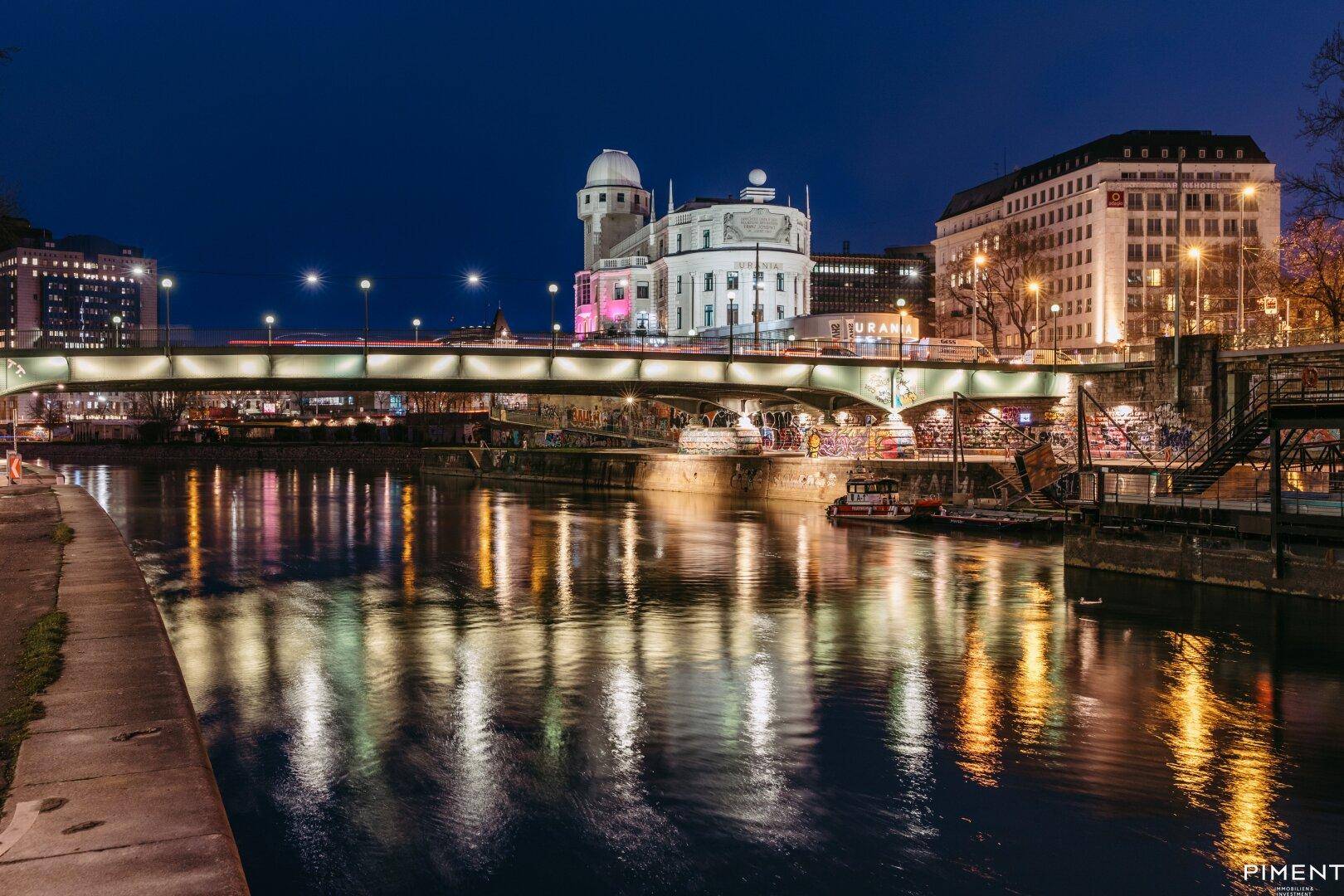 Nächtliche Stadtansicht mit beleuchteter Brücke, Fluss und historischen Gebäuden am Ufer.
