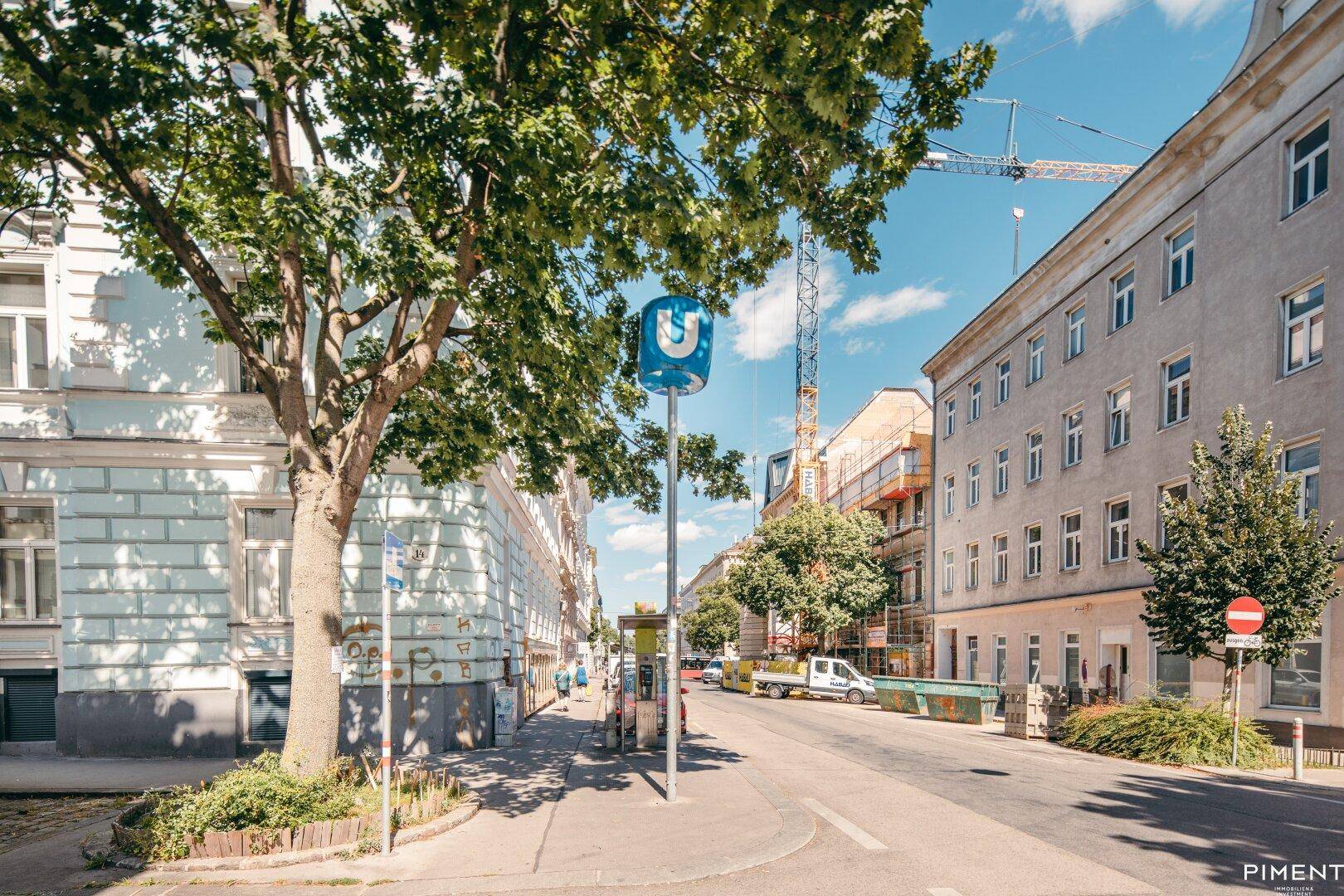 Belebtes Stadtbild mit U-Bahn-Haltestelle, Bäumen und umliegenden Gebäuden.