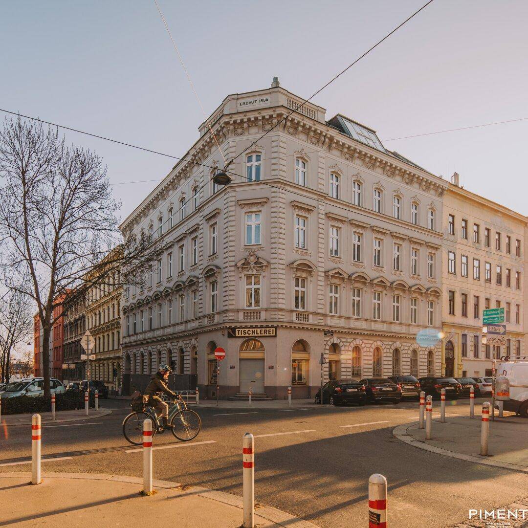 Historisches Eckgebäude an einer belebten Straße mit Radfahrer und parkenden Autos.