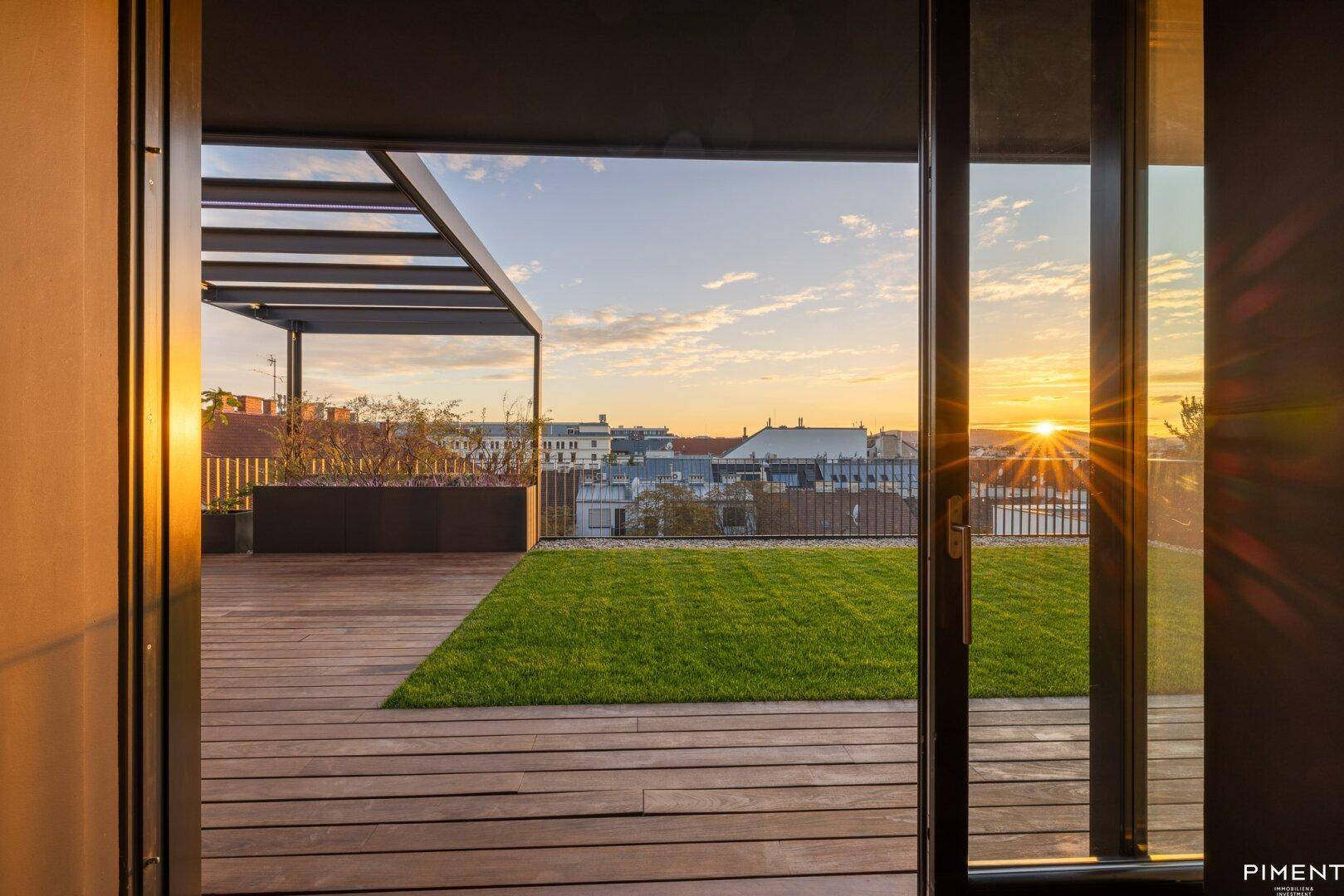 Panoramaaussicht von innen auf die Dachterrasse mit Rasenfläche und weitem Stadtblick bei Sonnenuntergang.