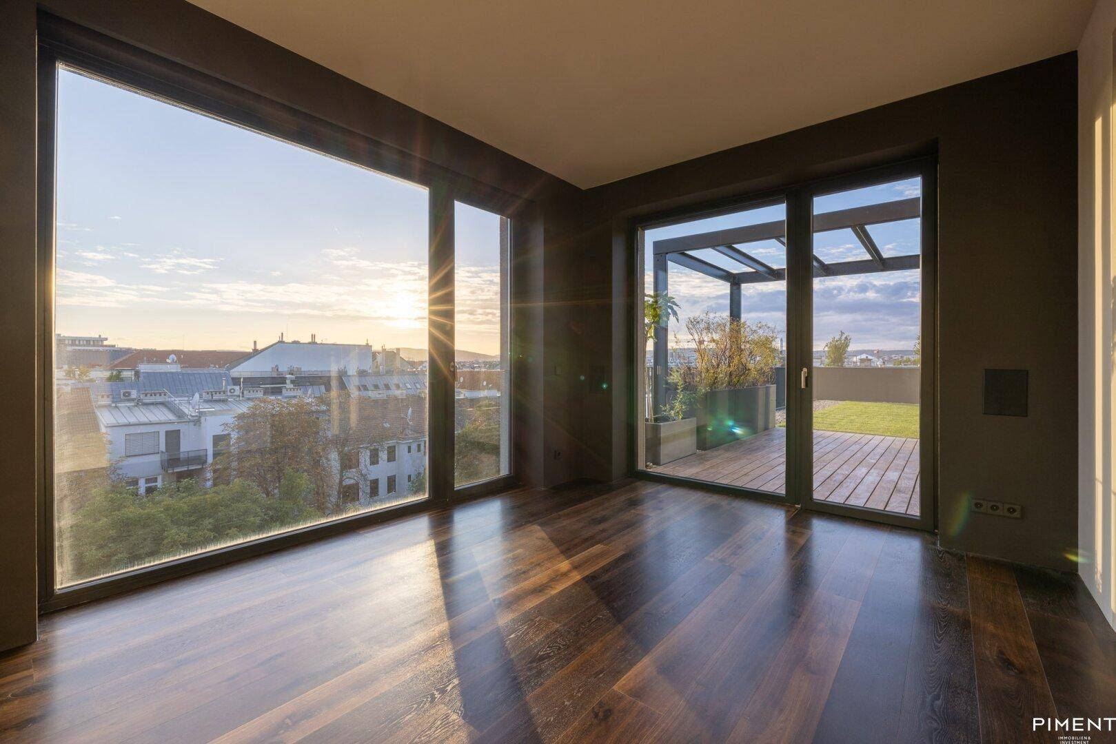 Geräumiger Raum mit dunklem Holzfußboden, großen Fenstern und Zugang zur Dachterrasse bei Sonnenuntergang.