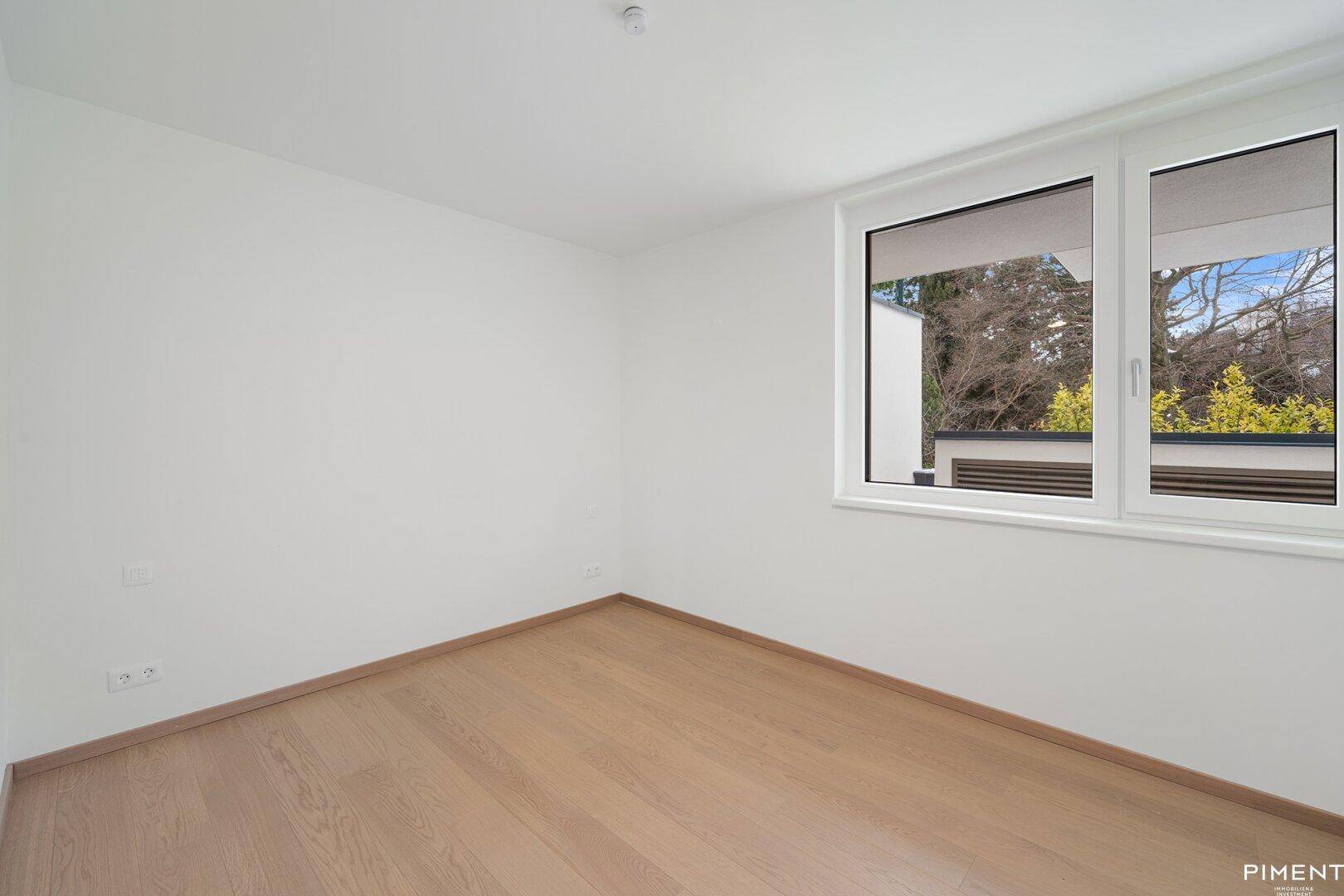 Helles, leeres Zimmer mit Holzfußboden und einem Fenster, das einen Blick ins Grüne ermöglicht.