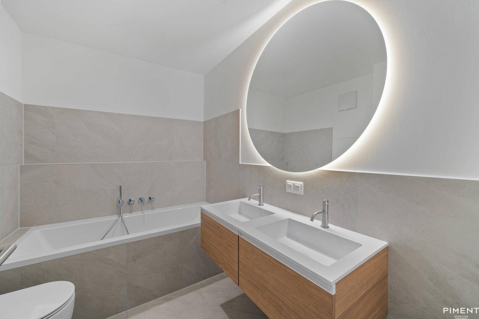 Modernes Badezimmer mit Badewanne, Doppelwaschbecken und einem großen, beleuchteten Spiegel über dem Waschtisch.