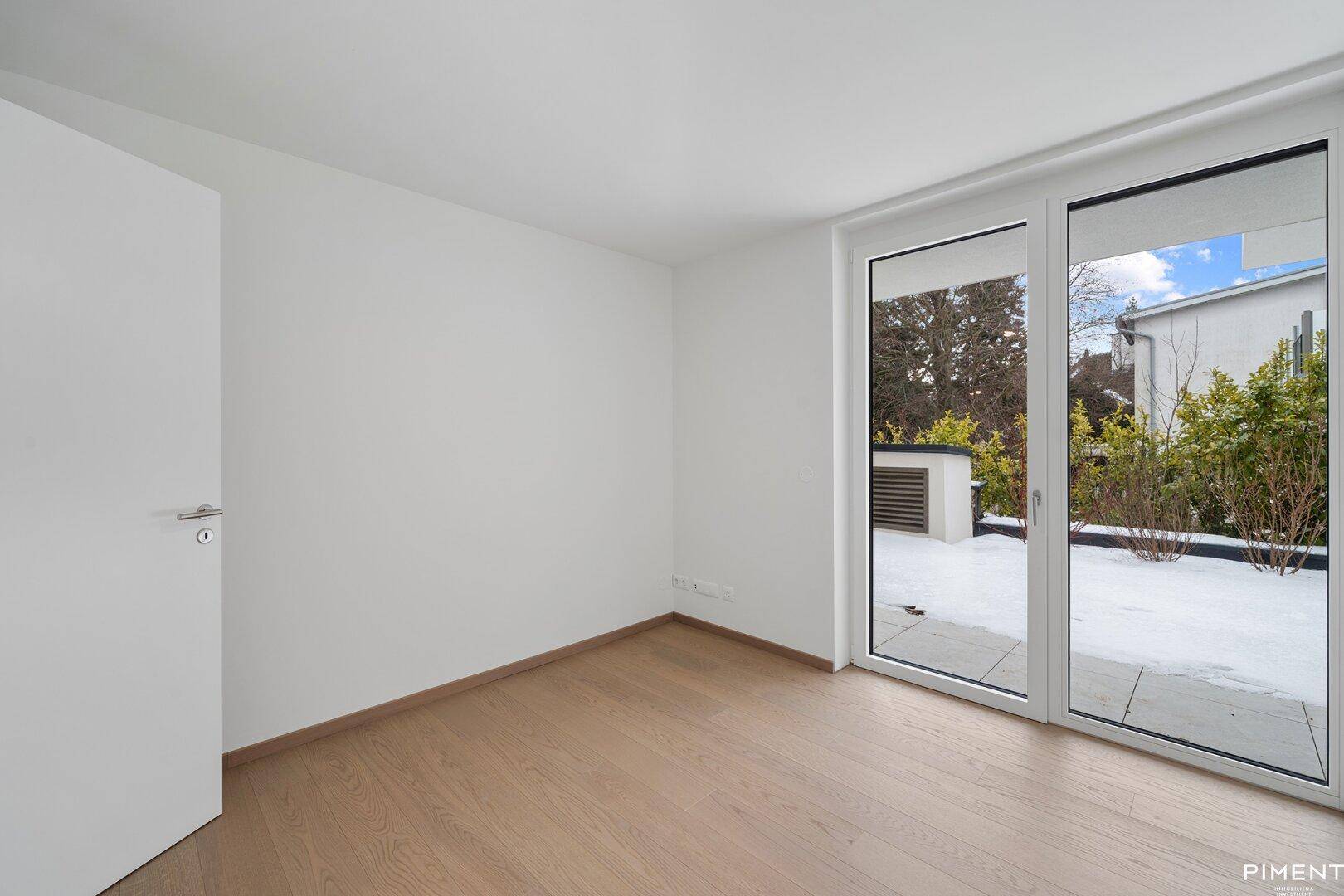 Helles, leeres Schlafzimmer mit hellem Holzfußboden und großen Fenstern, die Zugang zu einem Außenbereich bieten.