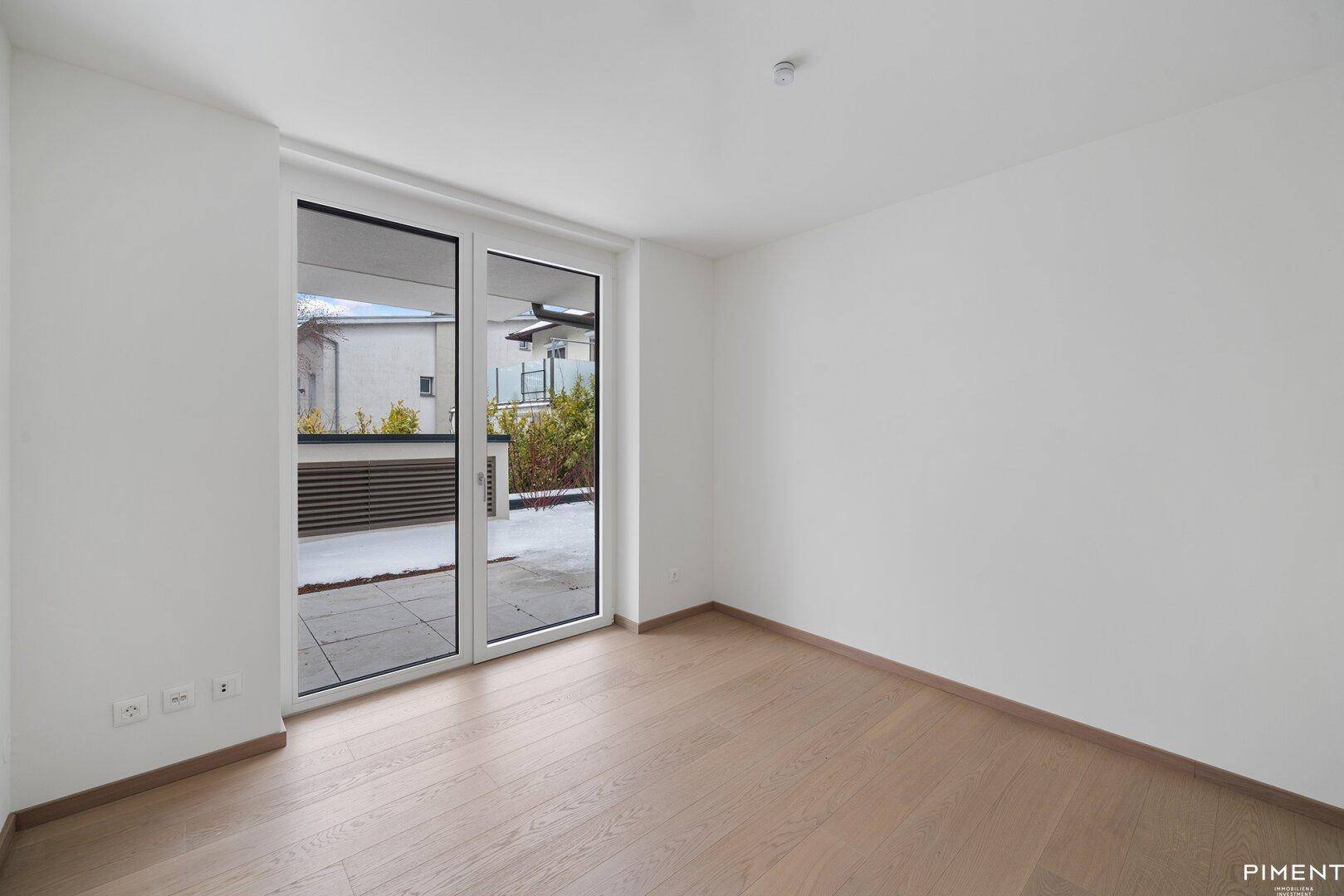 Helles, leeres Zimmer mit Holzboden und großen Fenstern, die Zugang zu einer Terrasse bieten.