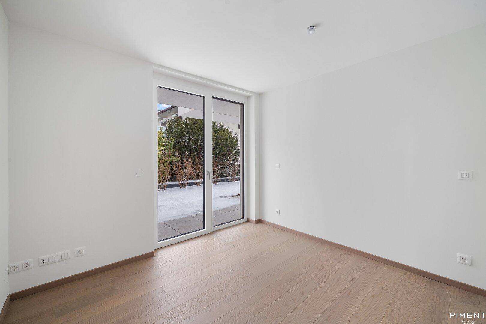 Helles, leeres Zimmer mit Holzboden und großen Fenstern, die Zugang zu einer Terrasse bieten.